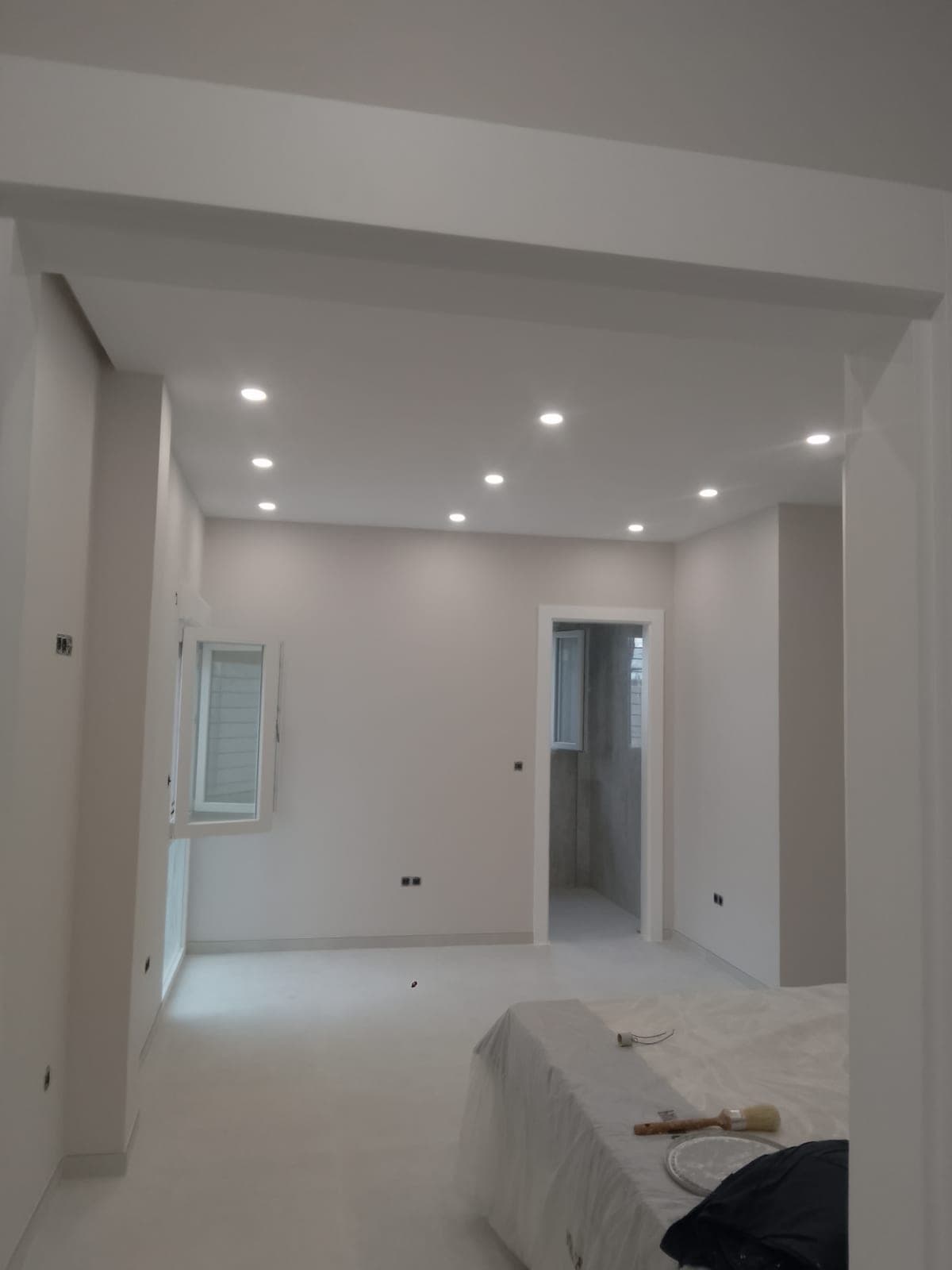 Dormitorio con el techo terminado y focos empotrados encendidos, junto a la puerta de un baño con las paredes alicatadas