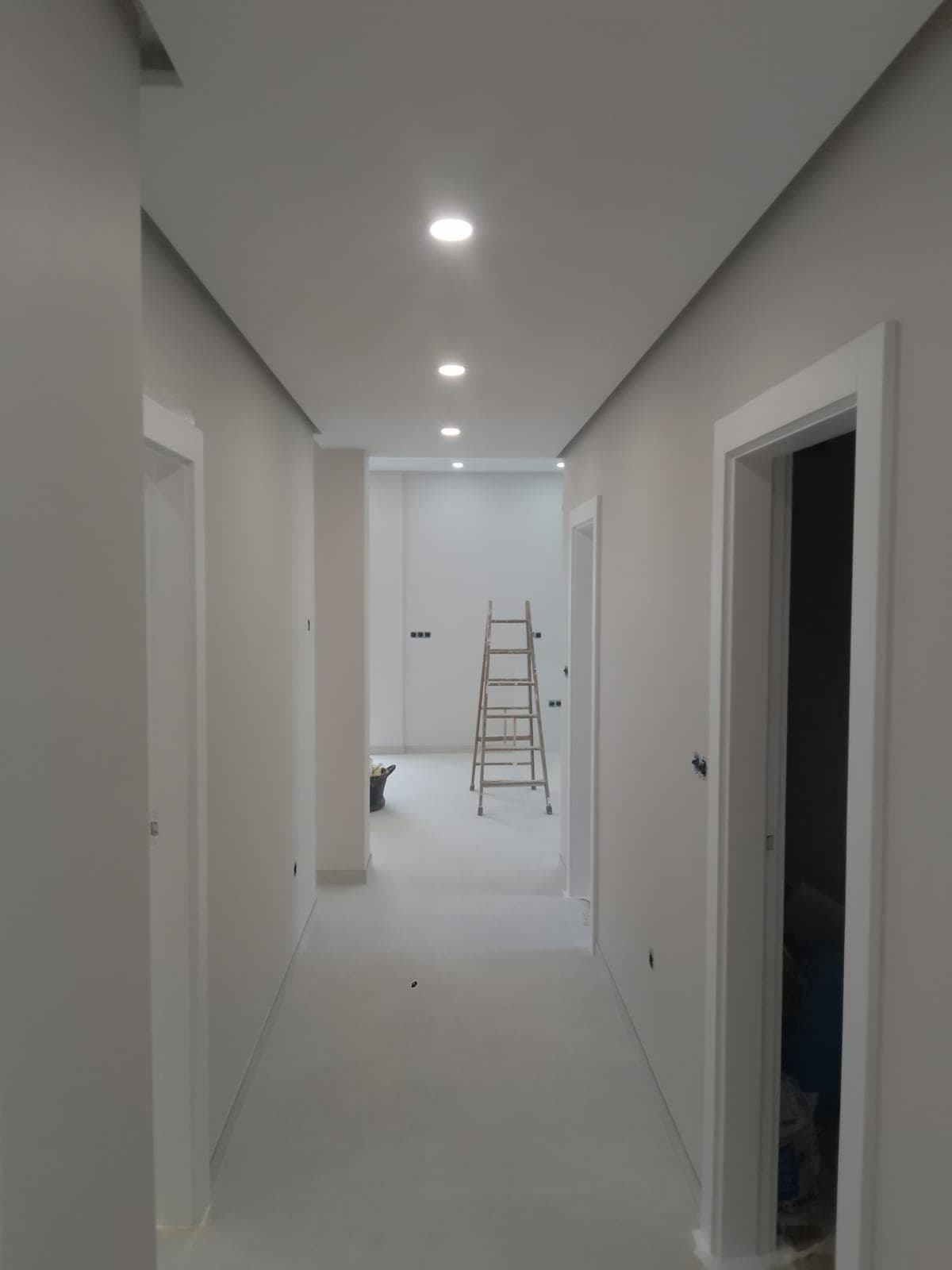 Tramo de pasillo con el techo terminado y focos empotrados, con una escalera de mano en la habitación del fondo
