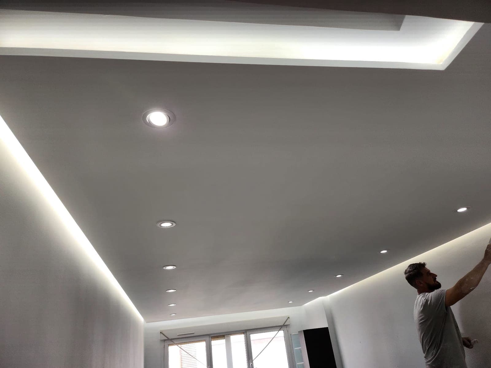 Vista amplia de un salón con el techo terminado y la iluminación indirecta encendida, mientras una persona continúa con los trabajos de pintura